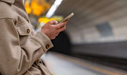 A passenger wearing a woven jacket is stood on an underground platform using their smart phone. 
